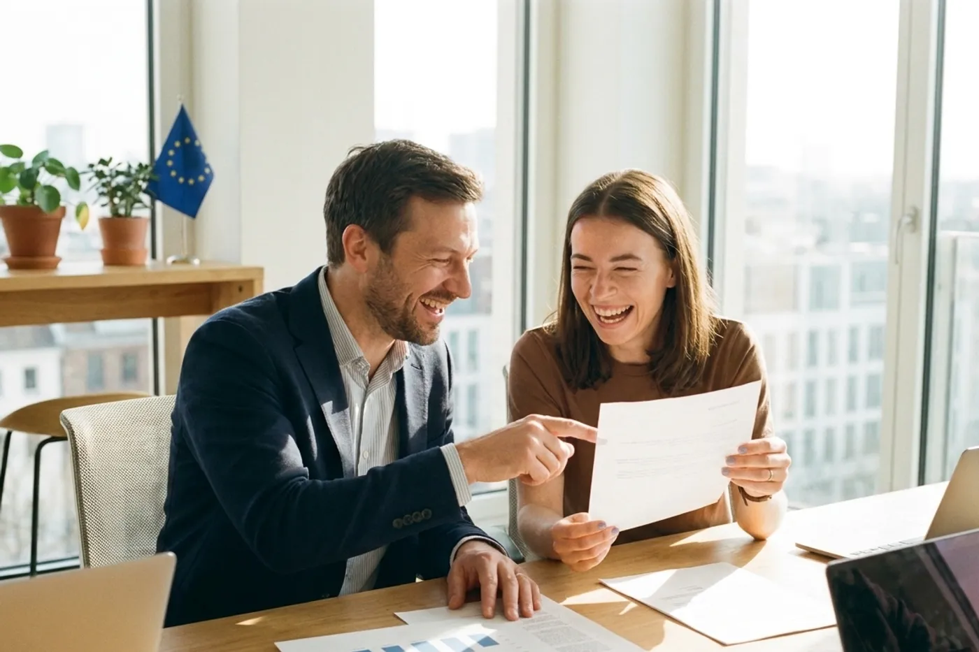 Two founders laughing over a funding decision letter at a desk with an EU flag behind them